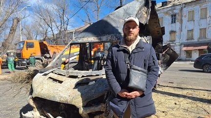 Casquette et barbe bien taillée, à 23 ans, David est employé municipal de la ville d'Odessa. Il pose devant une carcasse de voiture. (MARTIN TROADEC / FRANCEINFO / RADIO FRANCE)